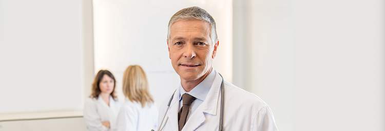 Un médecin, avec des cheveux gris et une blouse blanche, sourit.